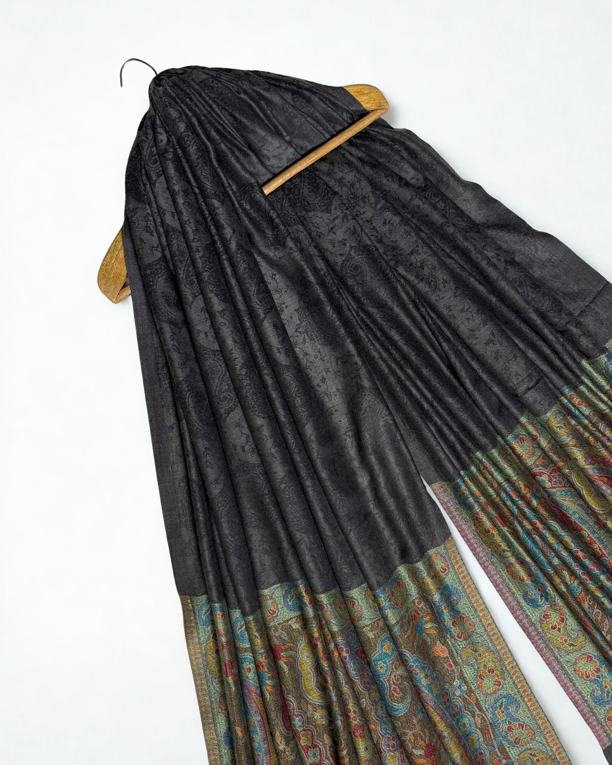 Dark charcoal fine pashmina ring shawl on a wooden hanger at an angle, showing the patterned field and woven paisley pallas