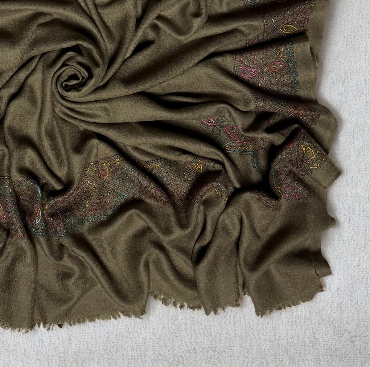 Close view of the olive brown Swati Shahtoosh shawl gathered in folds, the diamond lattice border worked in rose, teal, gold and pink