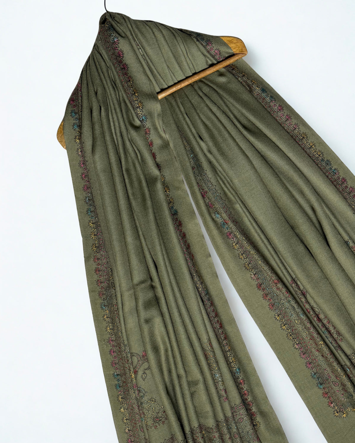 Olive green Swati Shahtoosh shawl draped over a wooden hanger, embroidered border through the folds