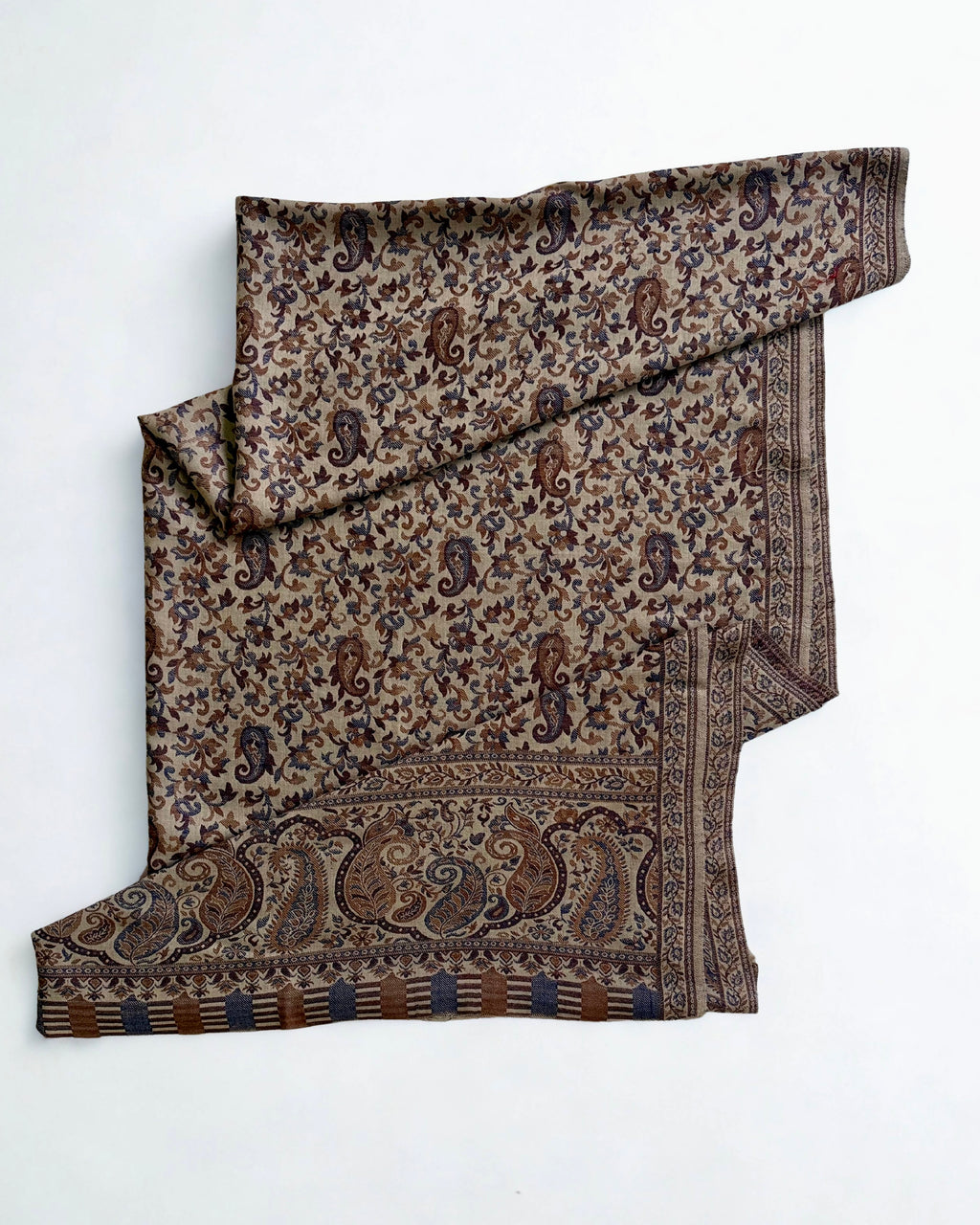 Sand-coloured Kani pashmina shawl laid flat, handwoven indigo and chestnut paisley field with a wide ornamented end border