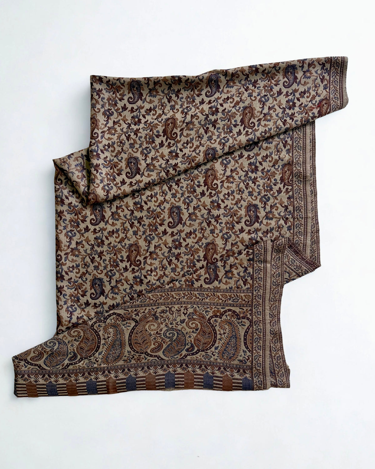 Sand-coloured Kani pashmina shawl laid flat, handwoven indigo and chestnut paisley field with a wide ornamented end border