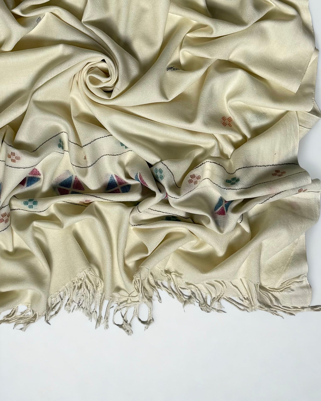 Close view of the off-white Khadi Swati Shahtoosh shawl in folds, showing the woven diamond medallion border in blue and rose and the twisted fringe