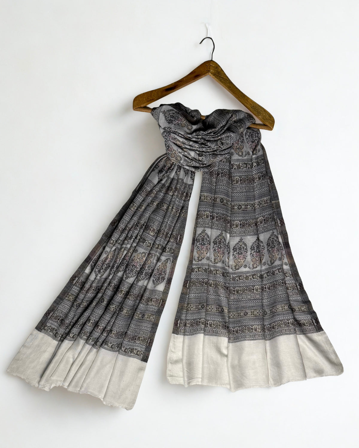 Silver grey Indian pashmina shawl on a wooden hanger, falling in fine pleats to show its full length and ivory end panels