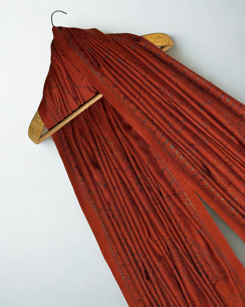 Rust Swati Shahtoosh shawl draped over a wooden hanger, embroidered border running through the folds