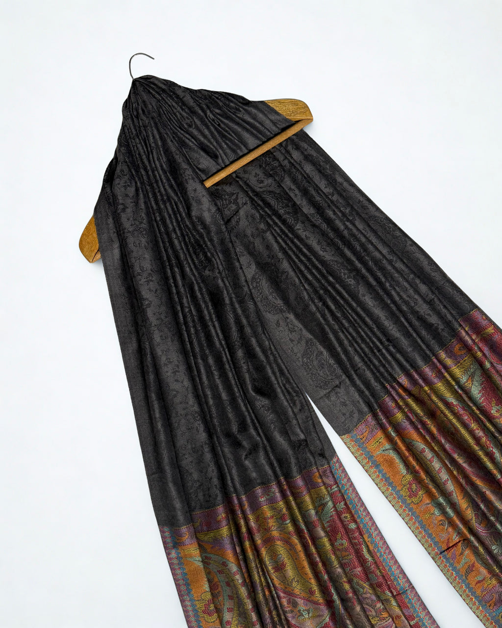Dark charcoal fine pashmina ring shawl on a wooden hanger at an angle, showing the patterned field and warm paisley pallas
